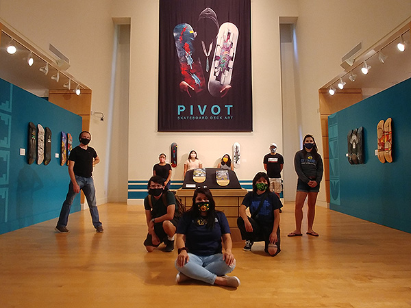 Eight student visitors from El Centro de Muchos Colores posing around the banner and the skateboard decks of the PIVOT: Skateboard Deck Art exhibit.